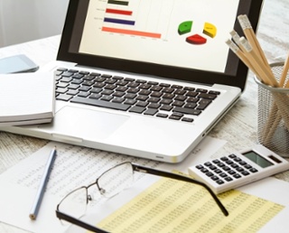 Cluttered desk with a laptop displaying charts, documents, eye glasses, and a calculator