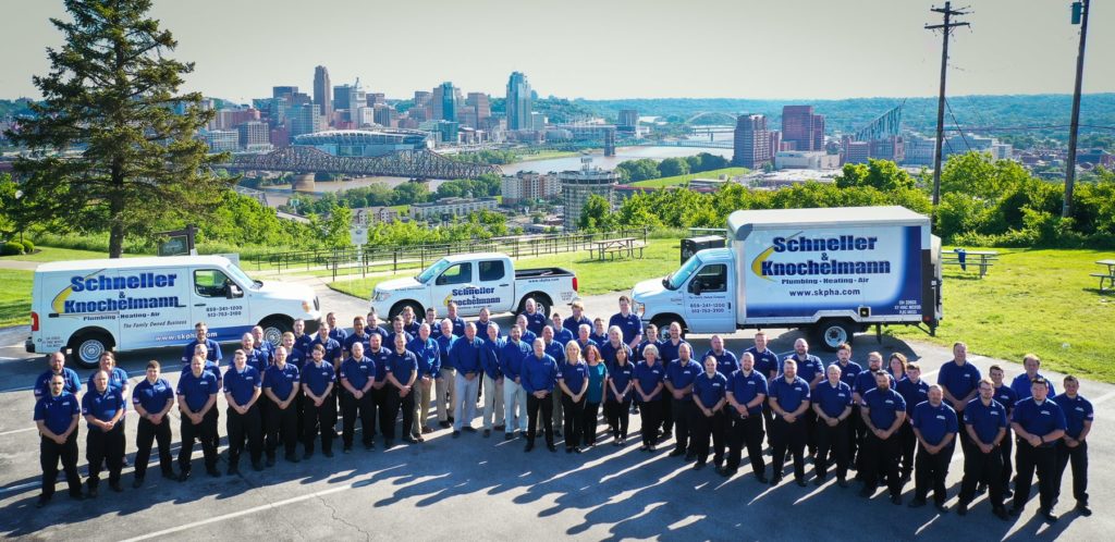 Schneller Knochelmann team photo. Aerial view of team standing together, all wearing blue polos and black pants, with service vehicles and city skyline in background.