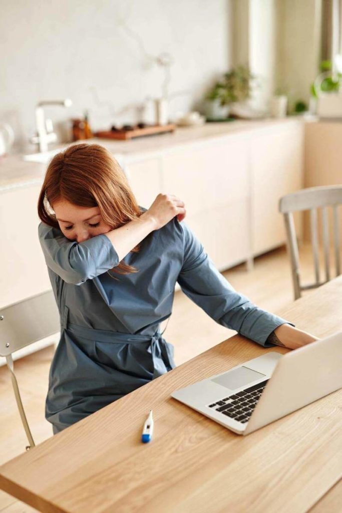 Young woman sneezing into arm while working on laptop at home.