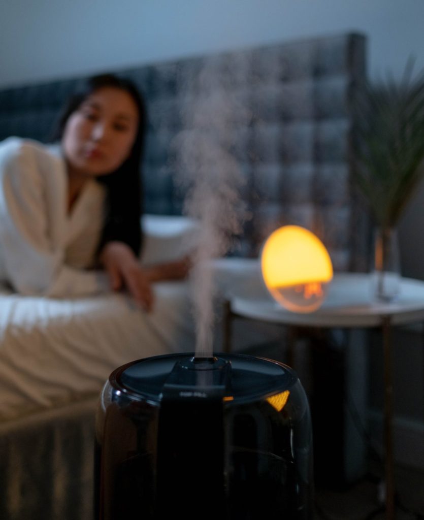 UV humidifier blowing vapor into the air; woman laying on bed in background.
