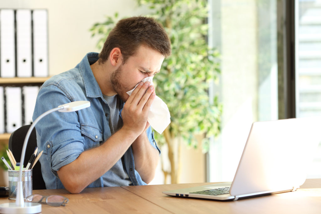 Side view of a young man sneezing in a wipe inside of his home office.