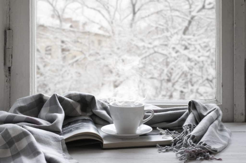 White cup of hot drink and opened book with gray and white plaid on white windowsill against snowy view of trees outside.