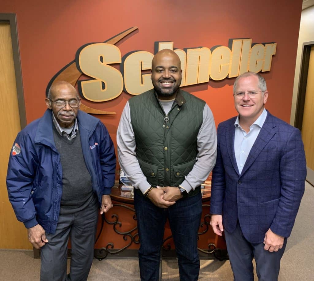 Three male men representing Schneller Knochelmann and Moore Air pose for photo in front of Schneller's company logo.