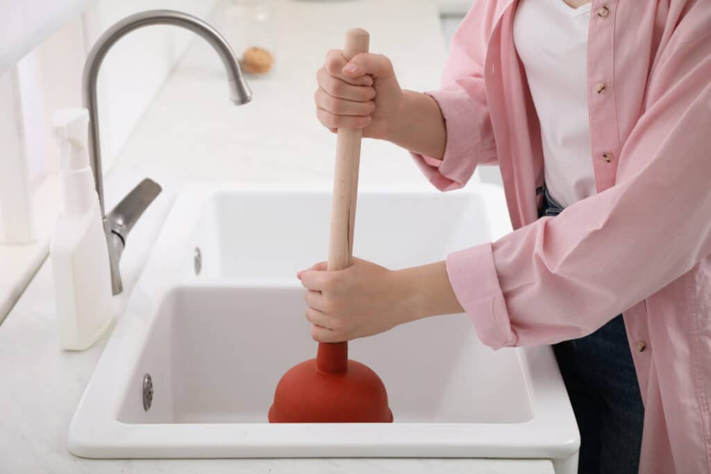 A person using a plunger to unclog the sink drain in their kitchen